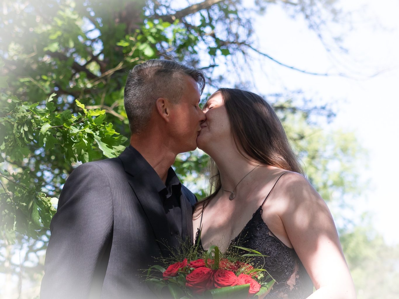 Een man en vrouw kussen elkaar, omringd door bomen en groen.