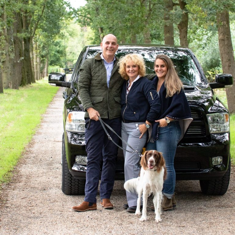 Drie mensen en een hond poseren naast een zwarte auto op een boomlanenweg.