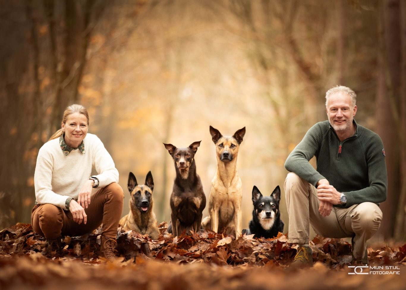 Een vrouw en man zitten op de grond tussen vier honden in een bosrijke omgeving. Foto gemaakt door Mijn Stijl Fotografie.